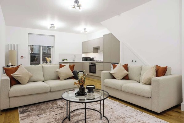 Ein modernes Wohnzimmer mit zwei Sofas, einem Glastisch und einer Küche im Hintergrund.