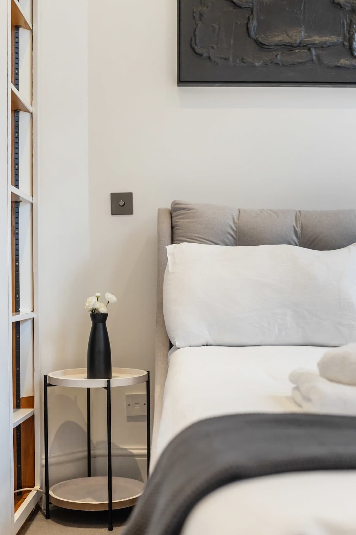 A bedroom with a bed, a bedside table with a vase, a bookshelf, and a wall-mounted light switch.