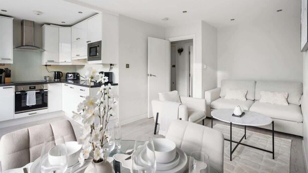 Una cocina moderna con armarios blancos, horno empotrado y microondas. Hay un comedor con una mesa y sillas blancas, y un sofá en la sala de estar.