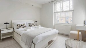 Un dormitorio moderno con una cama grande, mesitas de noche, una ventana con persianas y vistas a edificios.