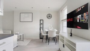 Una cocina moderna con armarios blancos, un comedor con una mesa redonda y sillas, un reloj de pared y un televisor de pantalla plana grande que muestra contenido de Netflix.