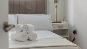 Ein ordentlich bezogenes Bett mit aufgerollten Handtüchern, einer Nachttischlampe und einem an der Wand hängenden Gemälde.