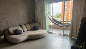 A living room with a sofa, a potted plant, and a view of a building through a glass door. - Apartment on Praia do Futuro beach, 200m from the sea. (Fortaleza)
