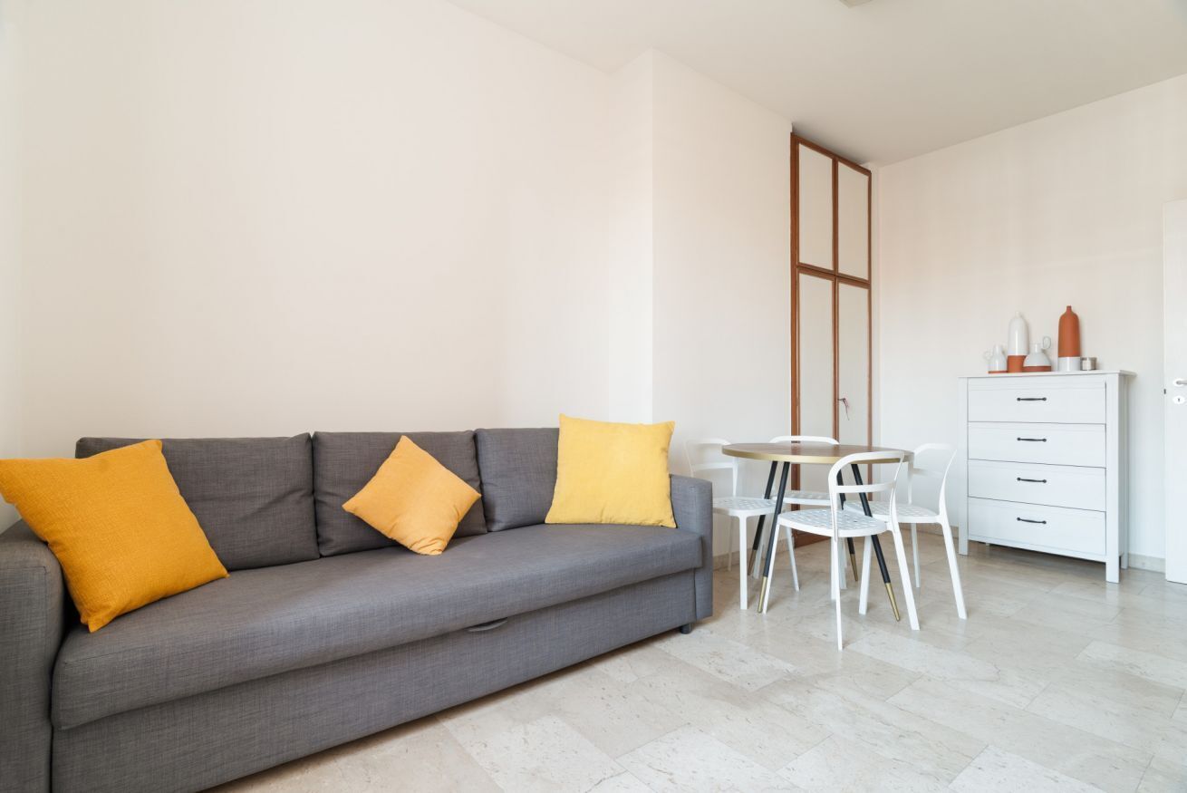 Un salon moderne avec un canapé gris, deux coussins jaunes, une table blanche avec des chaises et un meuble blanc.