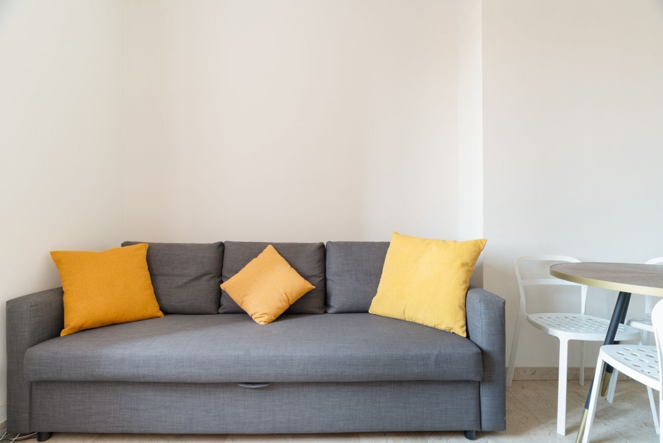 Un canapé gris avec quatre coussins, une petite table ronde avec des chaises et un mur blanc.