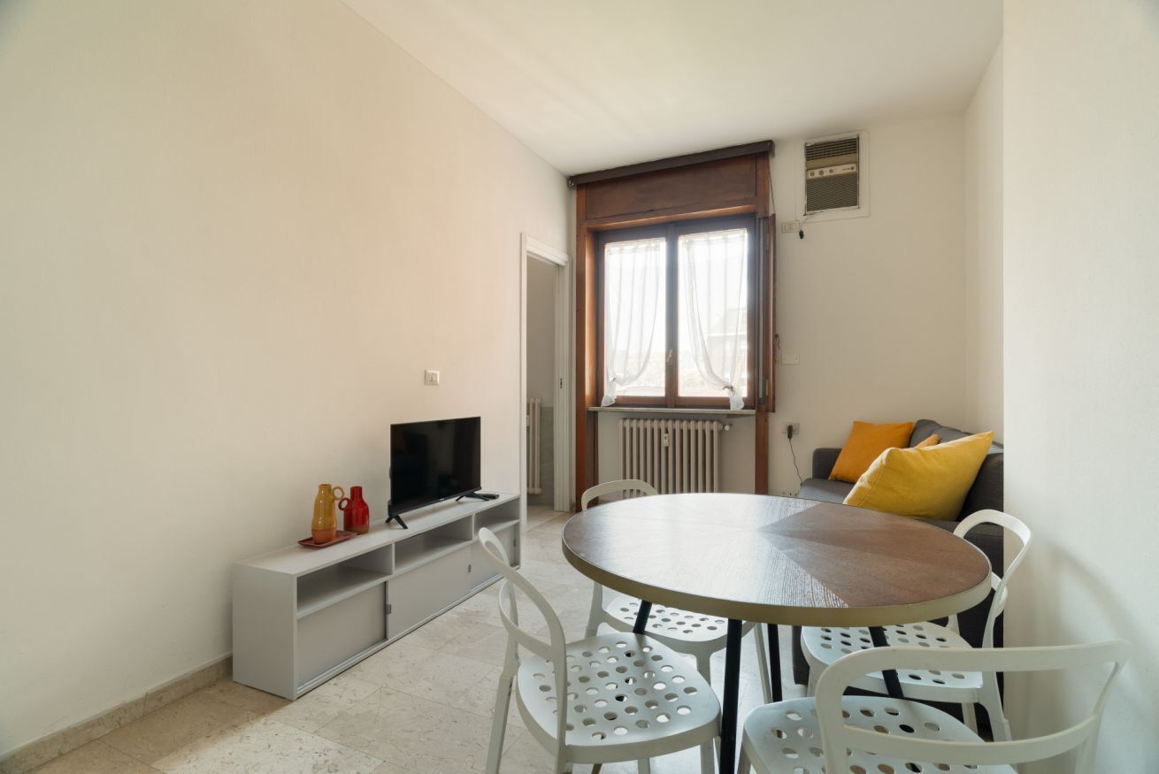 Une petite pièce bien éclairée, avec une table de salle à manger ronde, des chaises blanches, un téléviseur posé sur un meuble bas, un radiateur et une fenêtre avec des rideaux.