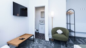 Ett modernt rum med en TV, ett träbord med fjärrkontroller, en grön fåtölj, en golvlampa och en klädställning.