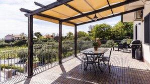 En overdækket terrasse med et bord, stole, grill og udsigt til grønne omgivelser.