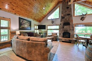 A cozy living room with a stone fireplace, wooden ceiling, and large windows. - Lake Namakagon Getaway for Boating, Fishing & Trail Riding (Cable)