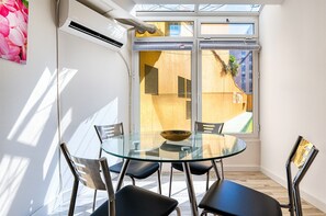 Ein moderner Essbereich mit einem Glastisch und Metallstühlen, einem großen Fenster mit Jalousien und Blick ins Freie.