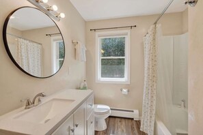 Un baño con un espejo redondo, un lavamanos, un inodoro, una ventana y una cortina de ducha.