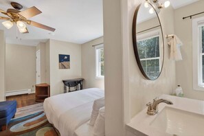 Un dormitorio con cama, ventilador de techo, mesita de noche, ventana y baño con lavabo y espejo.