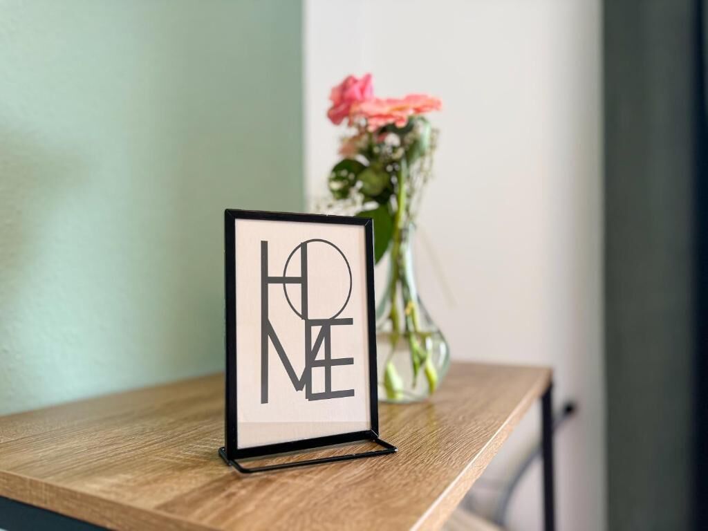 Et indrammet skilt med teksten 'HOME' på en træoverflade, en vase med blomster i baggrunden.