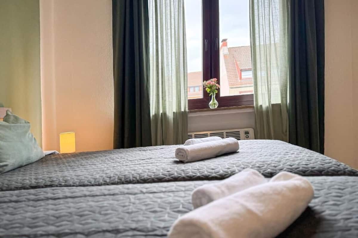 Ein Hotelzimmer mit Bett, einem Fenster mit Vorhängen und einer Vase mit Blumen auf dem Fensterbrett.