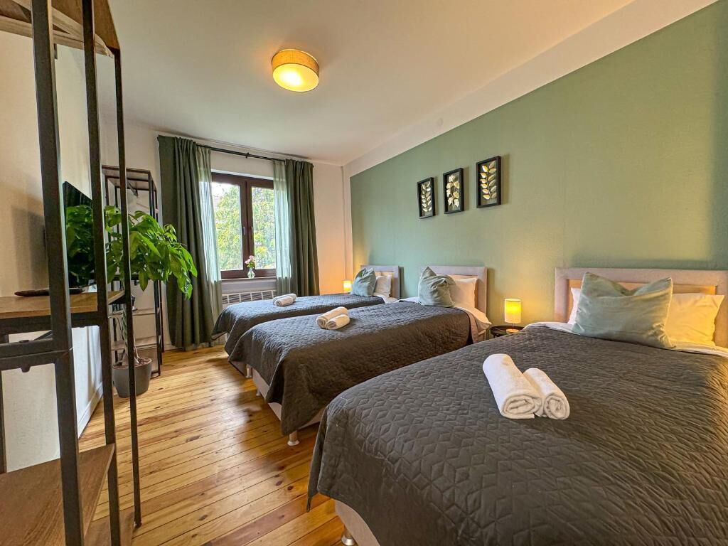 A room with two beds, a wooden floor, a window with curtains, and a plant.