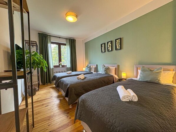 A room with two beds, a wooden floor, a window with curtains, and a plant.