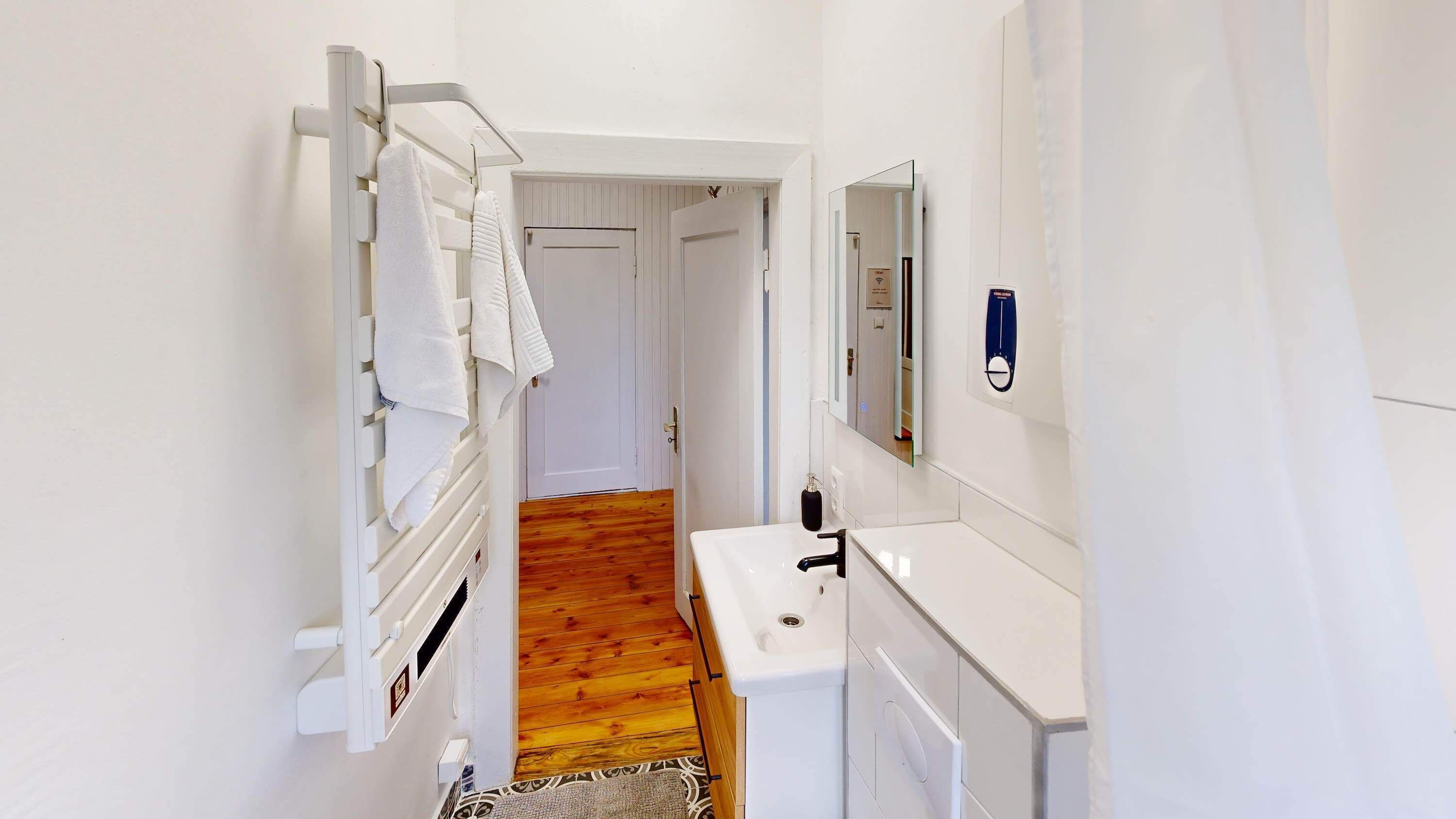 Een smalle gang met een witte radiator, een wit wastafelmeubel en een spiegel aan de muur.