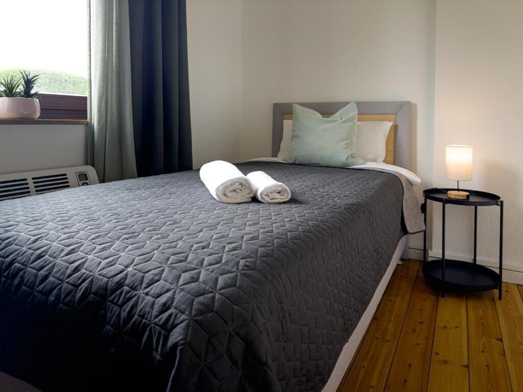A neatly made bed with a quilted bedspread, two rolled towels, a bedside lamp, and a small table with a lamp.