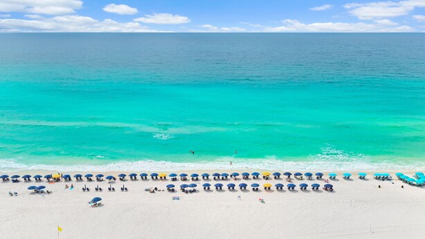 Une plage bordée de rangées de parasols et de chaises longues, un drapeau planté dans le sable, et l’océan en arrière-plan.