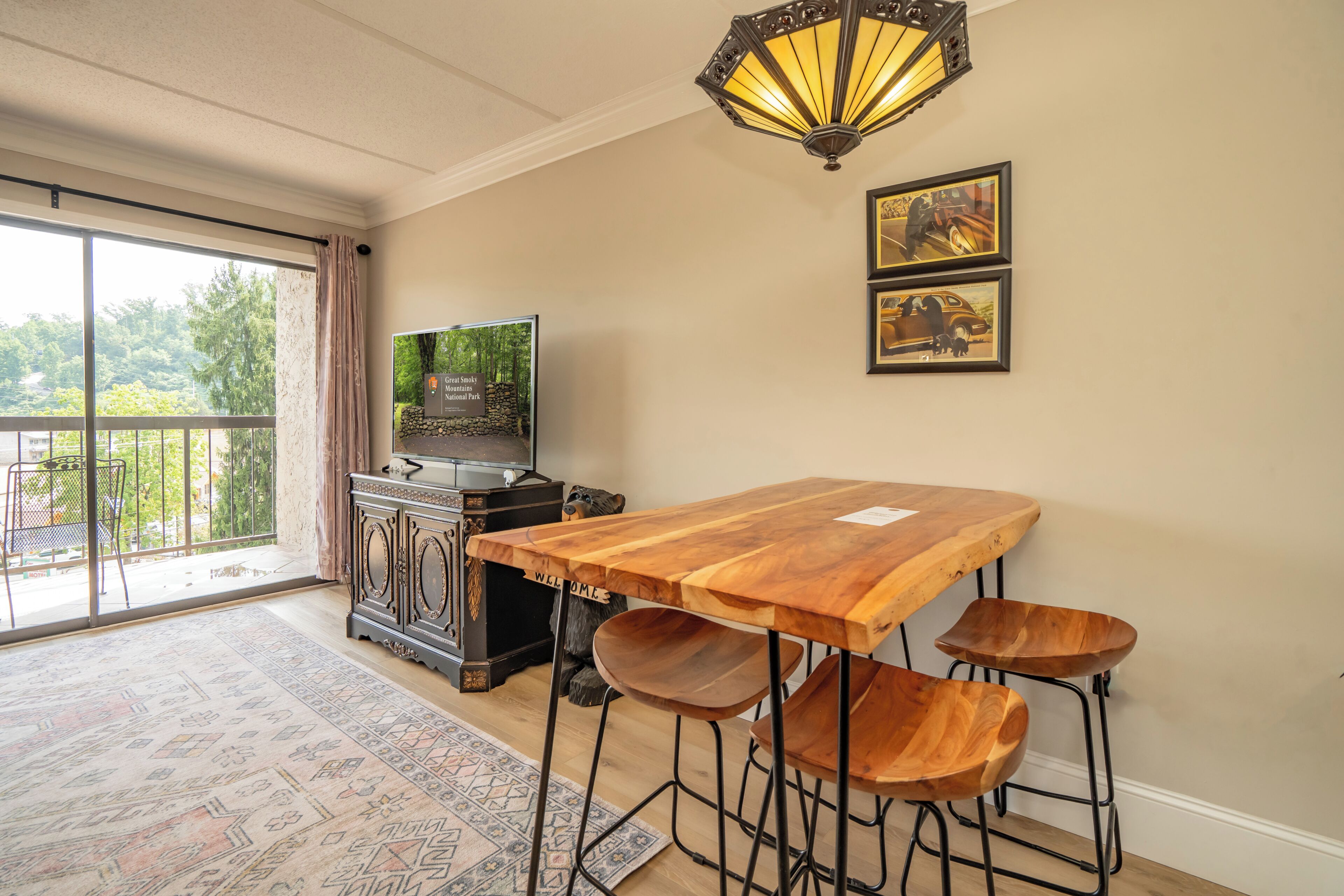 A dining area with a wooden table, bar stools, a television on a cabinet, and framed pictures on the wall.