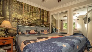 Un dormitorio con una cama con colcha de motivos forestales, una mesita de noche con lámpara y un espejo en la pared.