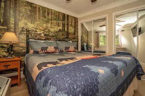 Een slaapkamer met een bed, voorzien van een quilt met een bosmotief, een nachtkastje met een lamp en een spiegel aan de muur.