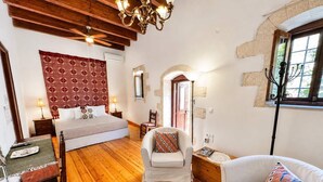 Una habitación con cama, sillón, sofá, piso de madera, pared con diseño y una lámpara de araña, además de una ventana.