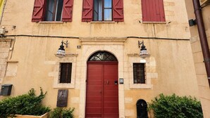 Un edificio con puerta roja, dos ventanas con contraventanas y un balcón pequeño.