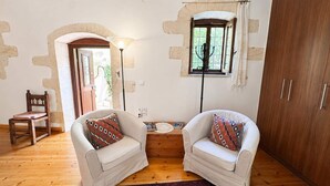 Una habitación con dos sillones blancos, suelo de madera, alfombra estampada, un armario de madera y una ventana con cortinas.