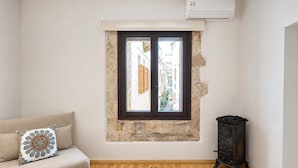 A room with a stone wall, a window with shutters, a sofa with a decorative pillow, a round wooden table, and a dark wooden cabinet. - Suite Victoria - Venetian Palace - Rooftop Jacuzzi (Chania)