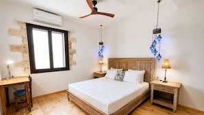 A bedroom with a wooden floor, a bed with a patterned headboard, two bedside tables with lamps, a ceiling fan, and a window with a view. - Suite Victoria - Venetian Palace - Rooftop Jacuzzi (Chania)