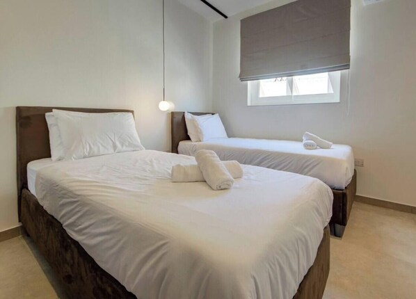 A hotel room with two beds, a window with a blind, and a hanging light fixture. - Fancy Sea View 3BR Apartment in Qawra | Sleeps 6 (San Pawl il-Baħar)