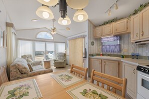 Un salon chaleureux avec un canapé, des fauteuils, une table à manger agrémentée de sets de table à motifs floraux, un ventilateur de plafond et un coin cuisine avec des placards et une cuisinière.