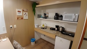 Uma cozinha compacta com micro-ondas, cafeteira e pia. Há prateleiras fixadas na parede acima do balcão.