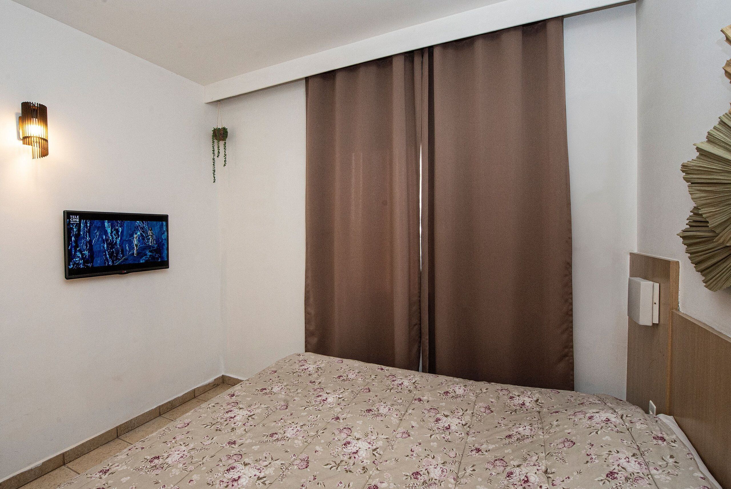 Una habitación de hotel con cama, televisor de pantalla plana, lámpara de pared y cortina.