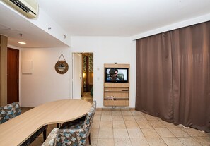 Una sala de estar con un televisor montado en la pared, una mesa de comedor con sillas estampadas y una cortina marrón que cubre una ventana.