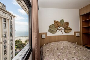 A hotel room with a bed, a wall-mounted decorative leaf, a window with a view of a beach and high-rise buildings, and a wooden shelf. - SHB - Family 2 Suítes Piscina e Academia (Santos)