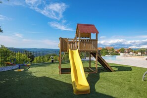 Ein Spielplatz mit Rutsche und Wippe, umgeben von einem Pool und Blick auf ferne Berge.