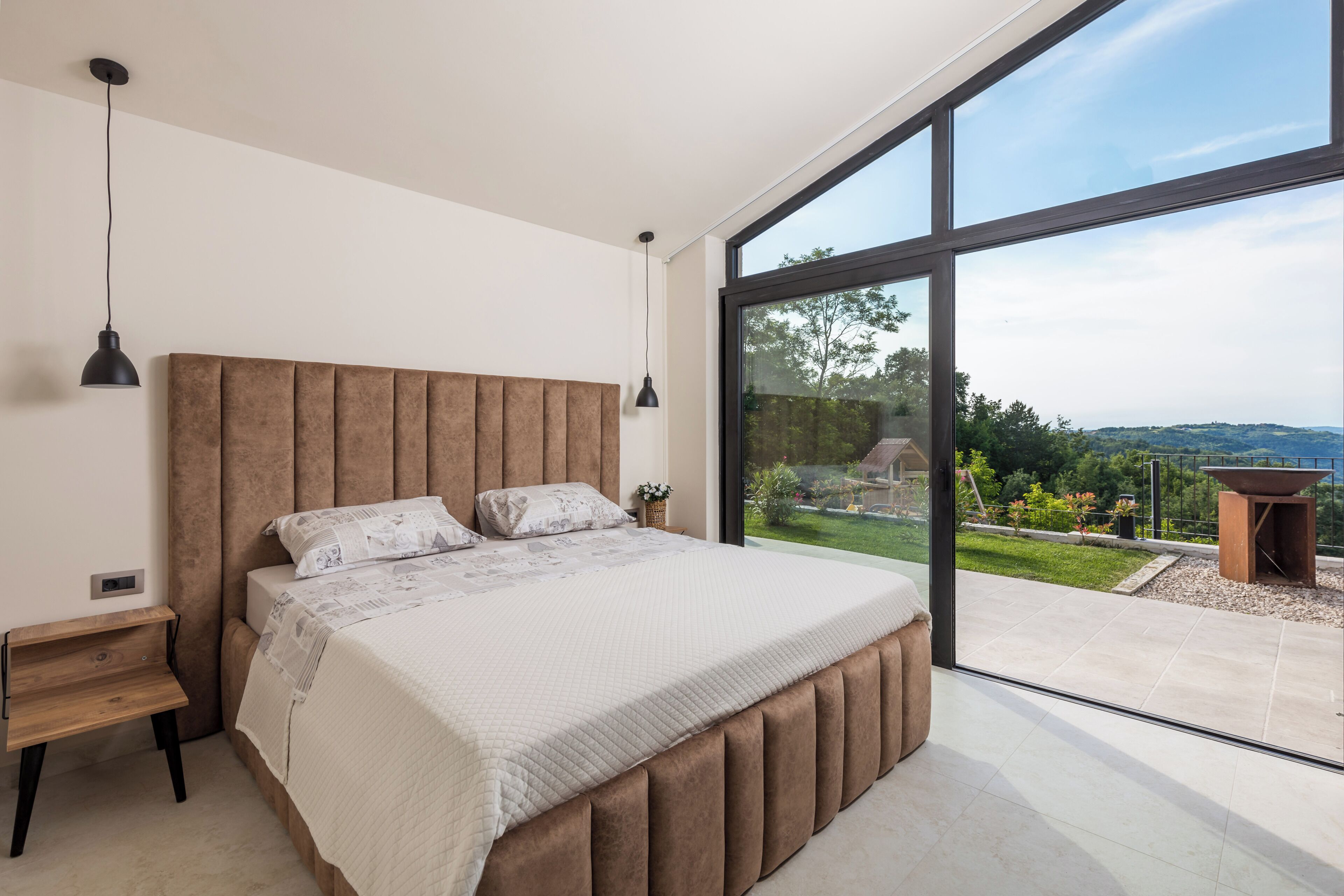 Une chambre moderne avec un grand lit, une table de chevet, une suspension et une vue sur un jardin, visible à travers une porte vitrée.