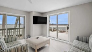 Una habitación amplia con un gran ventanal corredero que da a un balcón exterior, un televisor de pantalla plana montado en la pared y dos sofás a rayas.