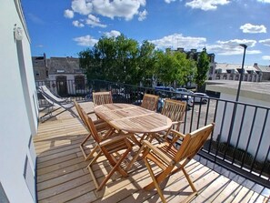 A wooden deck with a table and chairs, a railing, and a view of buildings and trees. - Ti ar Mor Bihan (Riantec)