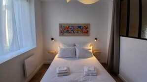 Een hotelkamer met een bed, witte gordijnen, een plafondlamp, een aan de muur hangend schilderij en bedlampjes.