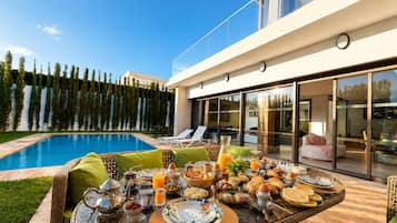 A modern outdoor dining area with a pool, wicker furniture, and a glass table set for breakfast.