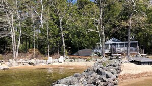 En stuga vid sjön med en brygga, omgiven av träd och en stenig strand.