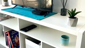 Een flatscreen-tv op een witte dressoir met een blauwe mat, een afstandsbediening, een toetsenbord, boeken, een kleine plant, een diffuser en een vaas.