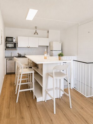 Eine moderne Küche mit weißen Schränken, einer zentralen Kochinsel, Barhocker, einer Mikrowelle, einem Kühlschrank und einer Deckenleuchte.