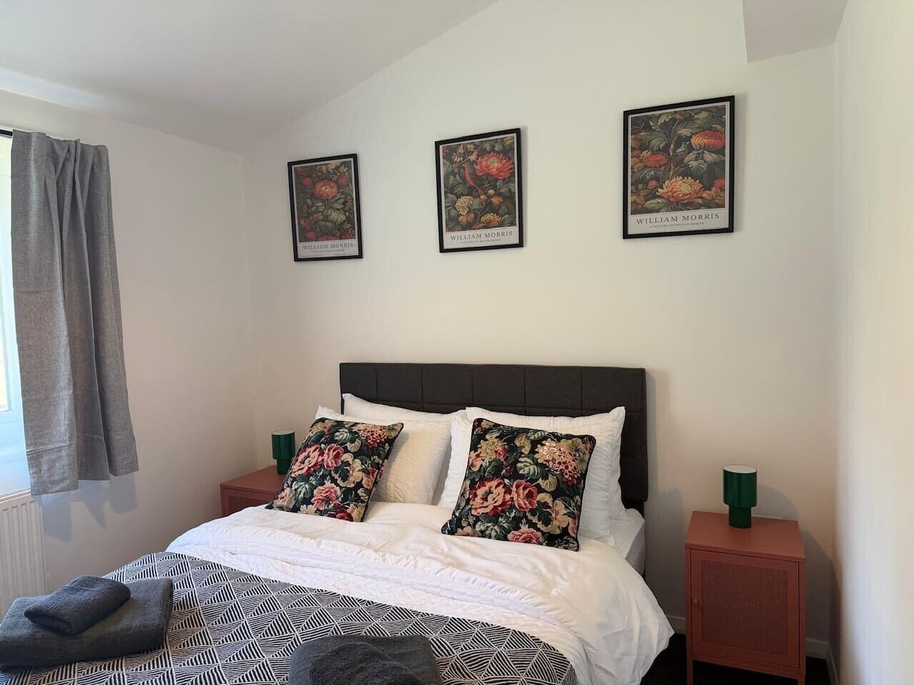 Une chambre à coucher avec un lit, des coussins à motifs floraux, une tête de lit, une table de chevet et une estampe botanique encadrée au mur.