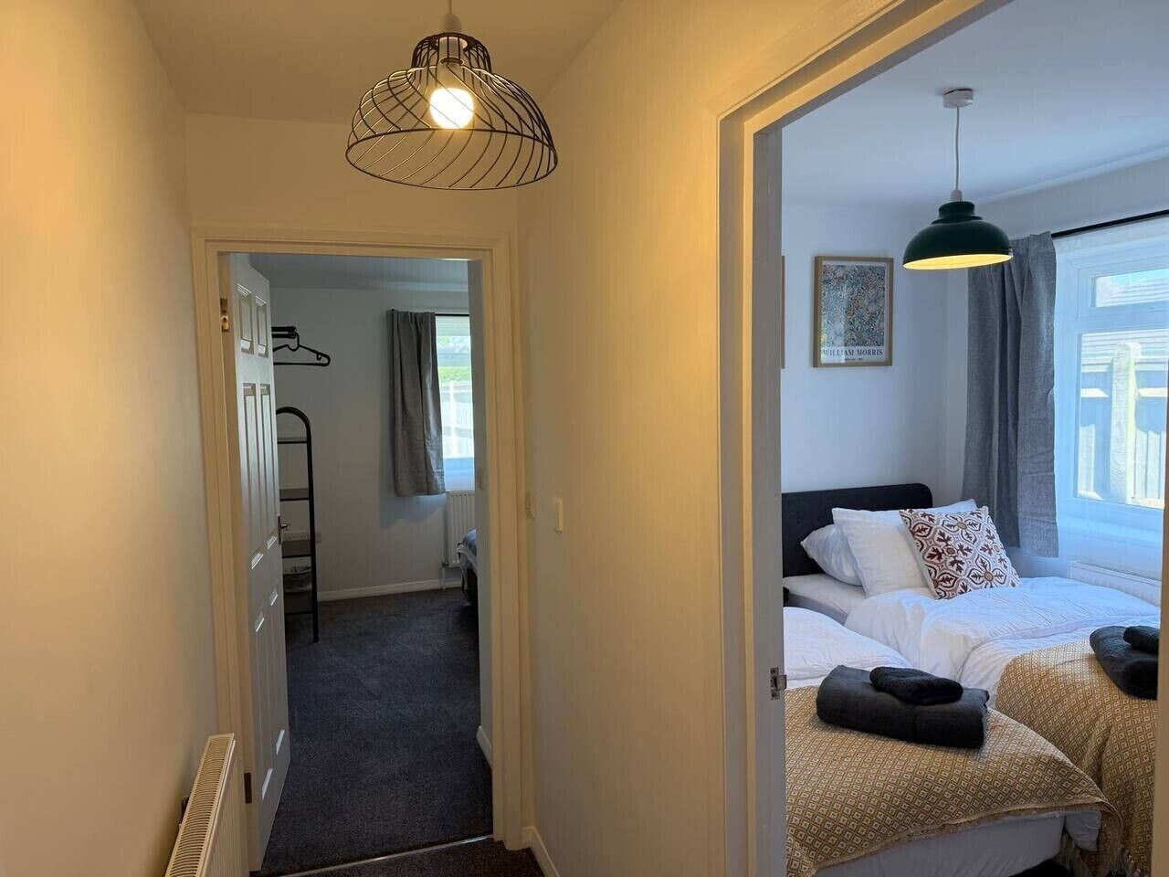 A hotel room with a bed, a small desk, a chair, a radiator, a window with curtains, and a hanging light fixture.