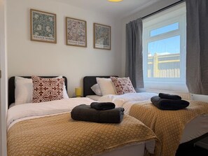 Ein Hotelzimmer mit zwei Betten, gemustertem Bettzeug, einem Fenster mit Vorhängen und drei gerahmten Kunstwerken an der Wand.