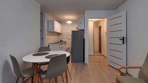 Une cuisine compacte dotée d’appareils électroménagers modernes, un coin repas avec une table blanche et des chaises grises, et un couloir menant à une autre pièce.